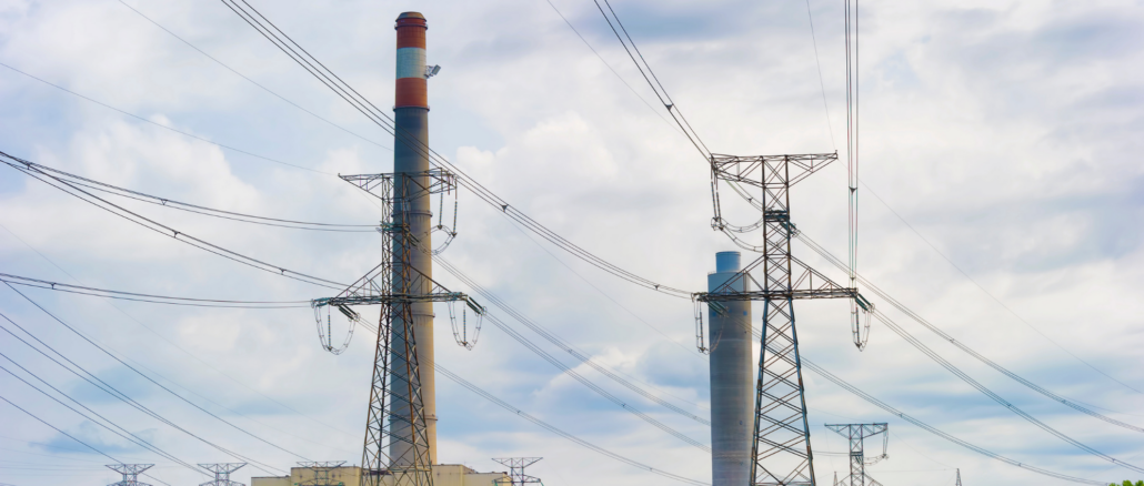 Industrial power plant with tall smokestacks and a lattice transmission tower amid a web of power lines against a cloudy sky.