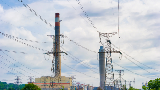 Industrial power plant with tall smokestacks and a lattice transmission tower amid a web of power lines against a cloudy sky.